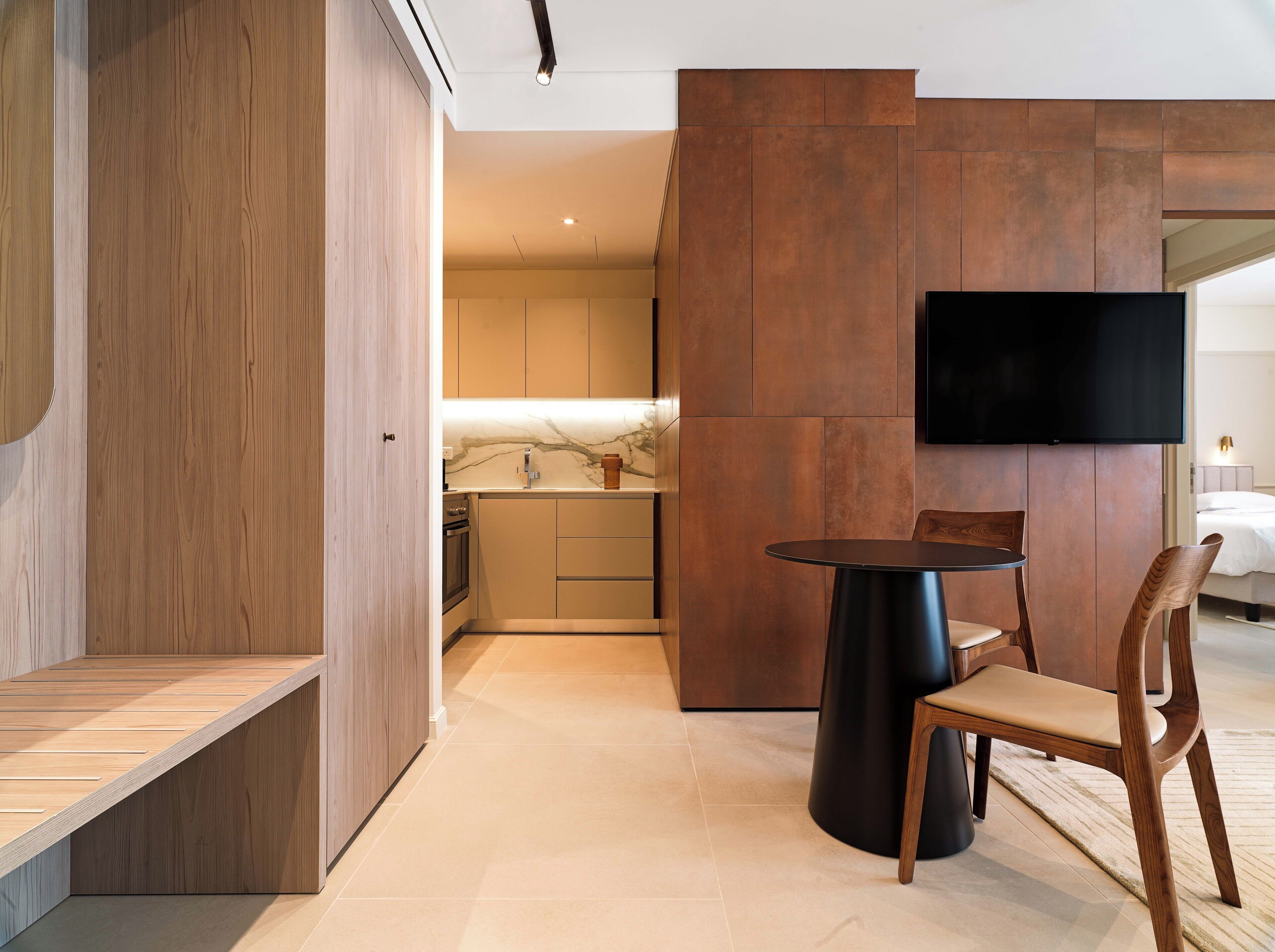 Ein modernes Hotelzimmer mit einer hölzernen Sitzbank, einem runden Tisch, einem Stuhl, einem flachbildfernseher an der Wand und einer Küchenzeile im Hintergrund.