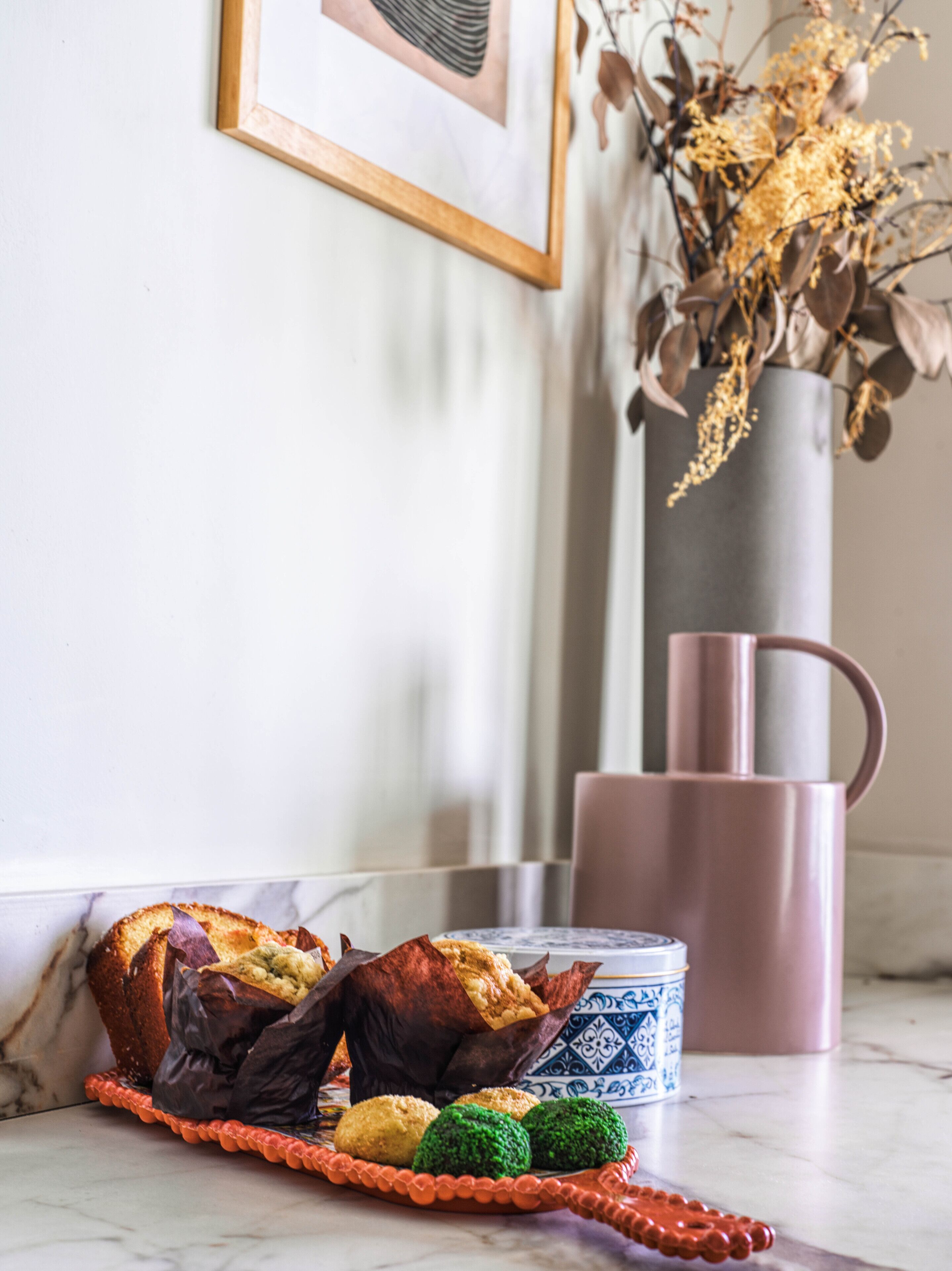 A kitchen counter with a plate of muffins, a vase with dried flowers, and a pink mug.