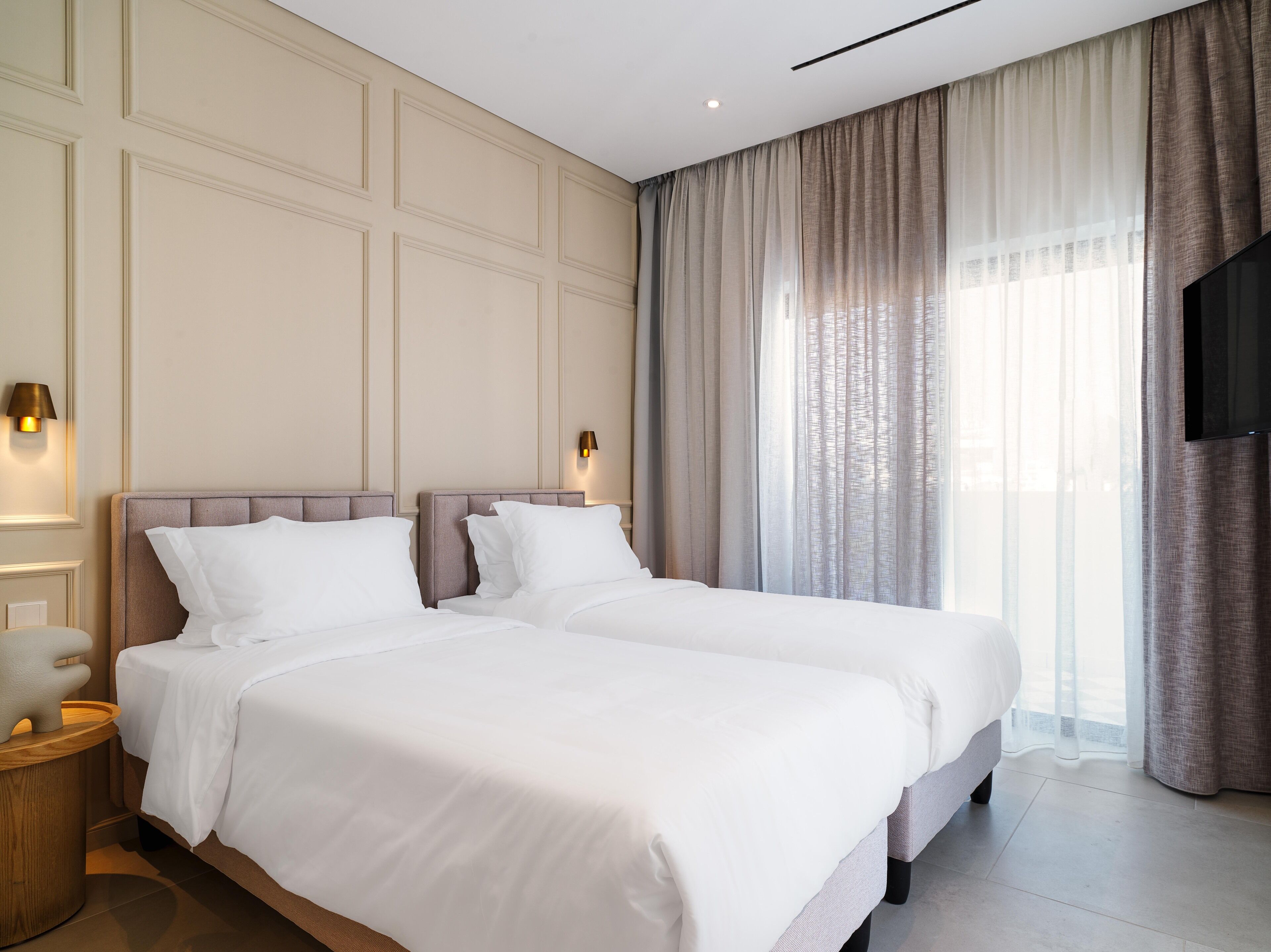 A hotel room with two beds, a flat-screen TV, and a large window with sheer curtains.