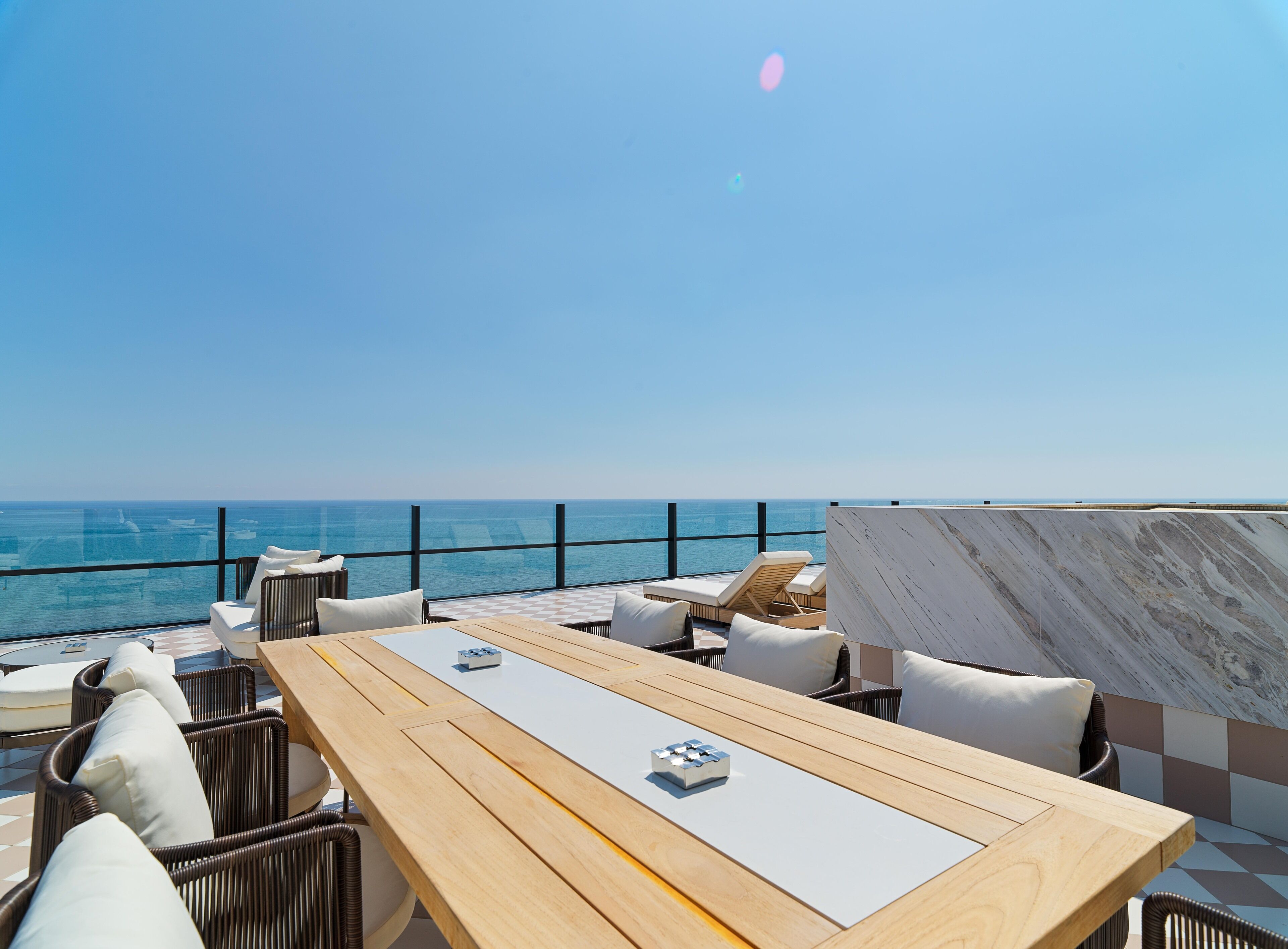 A rooftop terrace with a view of the ocean, furnished with wooden tables and wicker chairs.