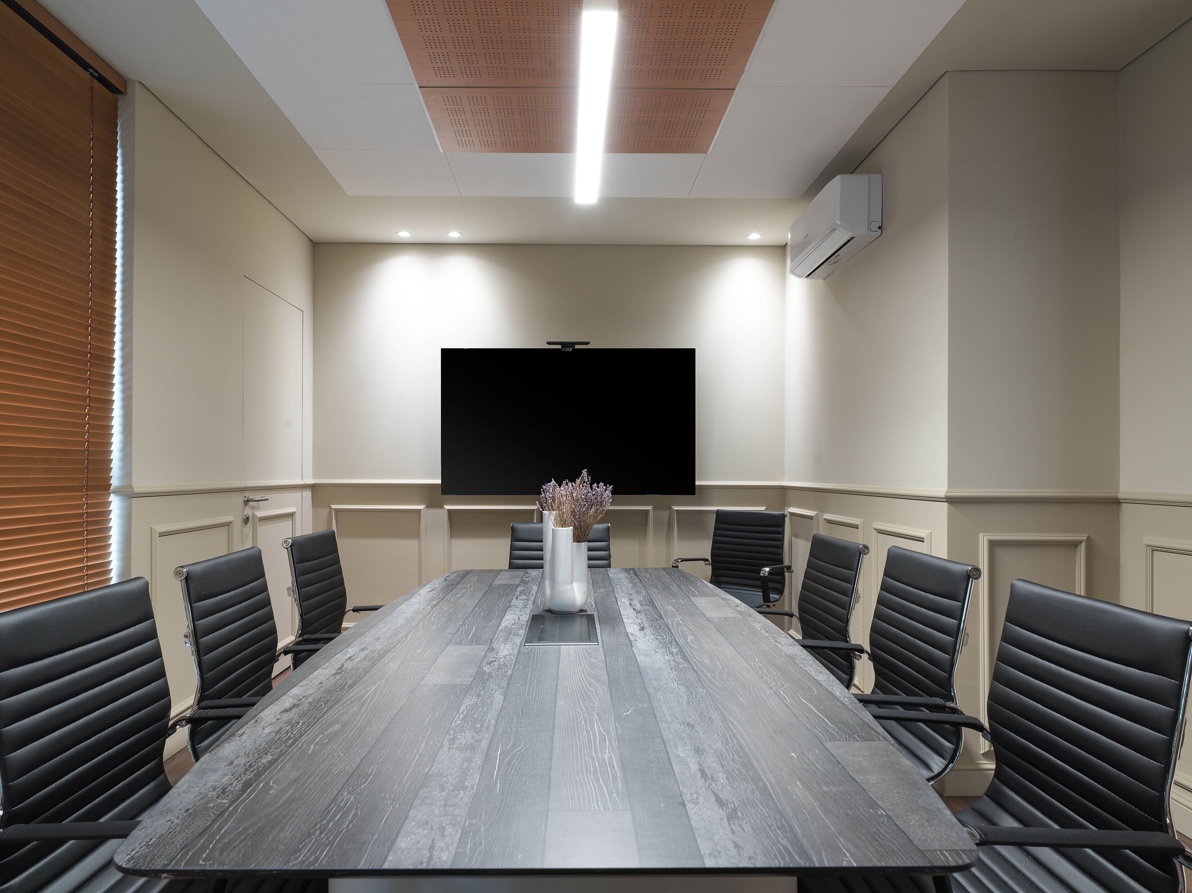 A conference room with a long table, black leather chairs, a large flat-screen TV, and a ceiling light fixture.