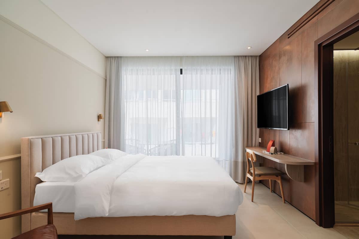 Una habitación de hotel con una cama grande, un escritorio con silla, un televisor de pantalla plana y un ventanal con cortinas translúcidas.