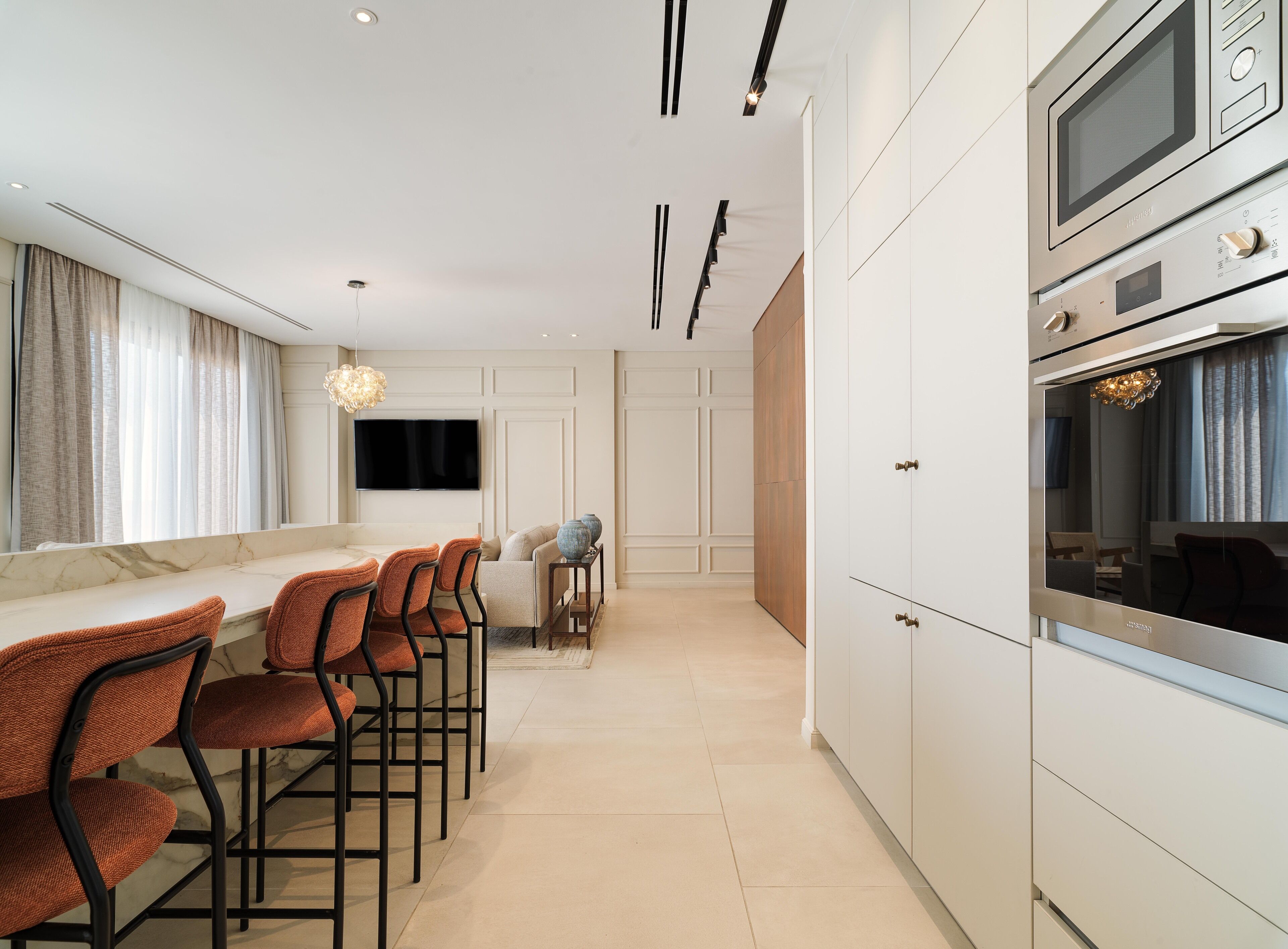 A modern kitchen with white cabinetry, a built-in oven, and a large TV. There are orange chairs and a dining table with a vase on it.