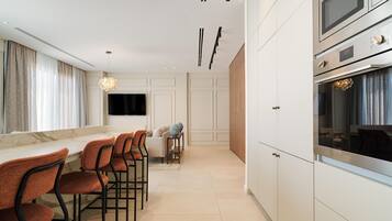 A modern kitchen with white cabinetry, a built-in oven, and a large TV. There are orange chairs and a dining table with a vase on it.
