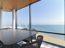 Ein moderner Essbereich mit großem Tisch und Korbstühlen, mit Blick auf einen Strand und den Ozean.