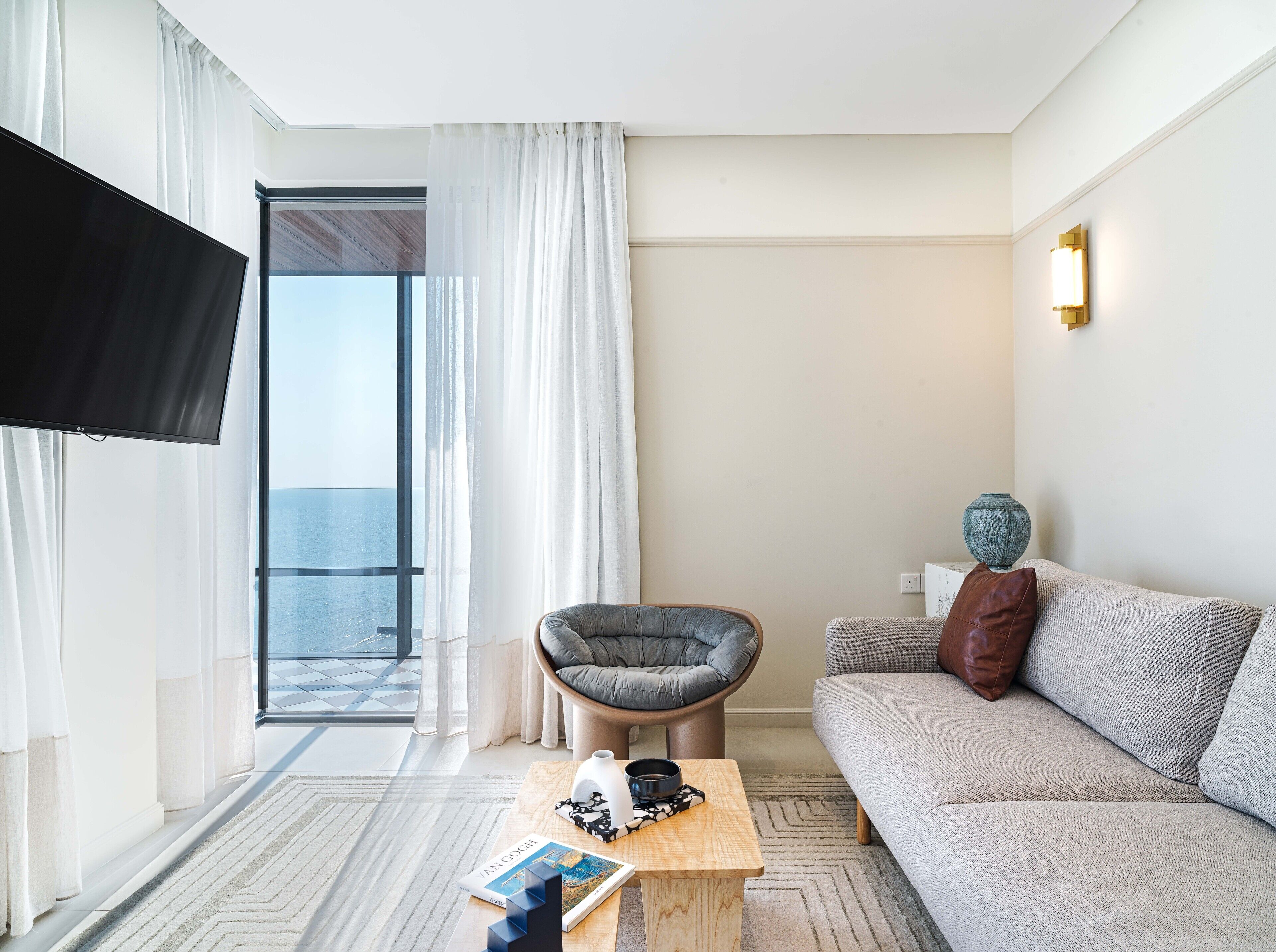 A modern hotel room with a sofa, a chair, a coffee table with a book, a vase, and a TV.