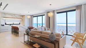 A modern living room with a sofa, coffee table, and bar stools. A large window offers a view of the sea.