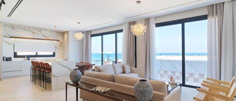 Un salon moderne avec un canapé, une table basse et des tabourets de bar. Une grande fenêtre offre une vue sur la mer.