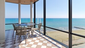 A balcony with a checkered floor, a table, and chairs, overlooking a beach and the sea.