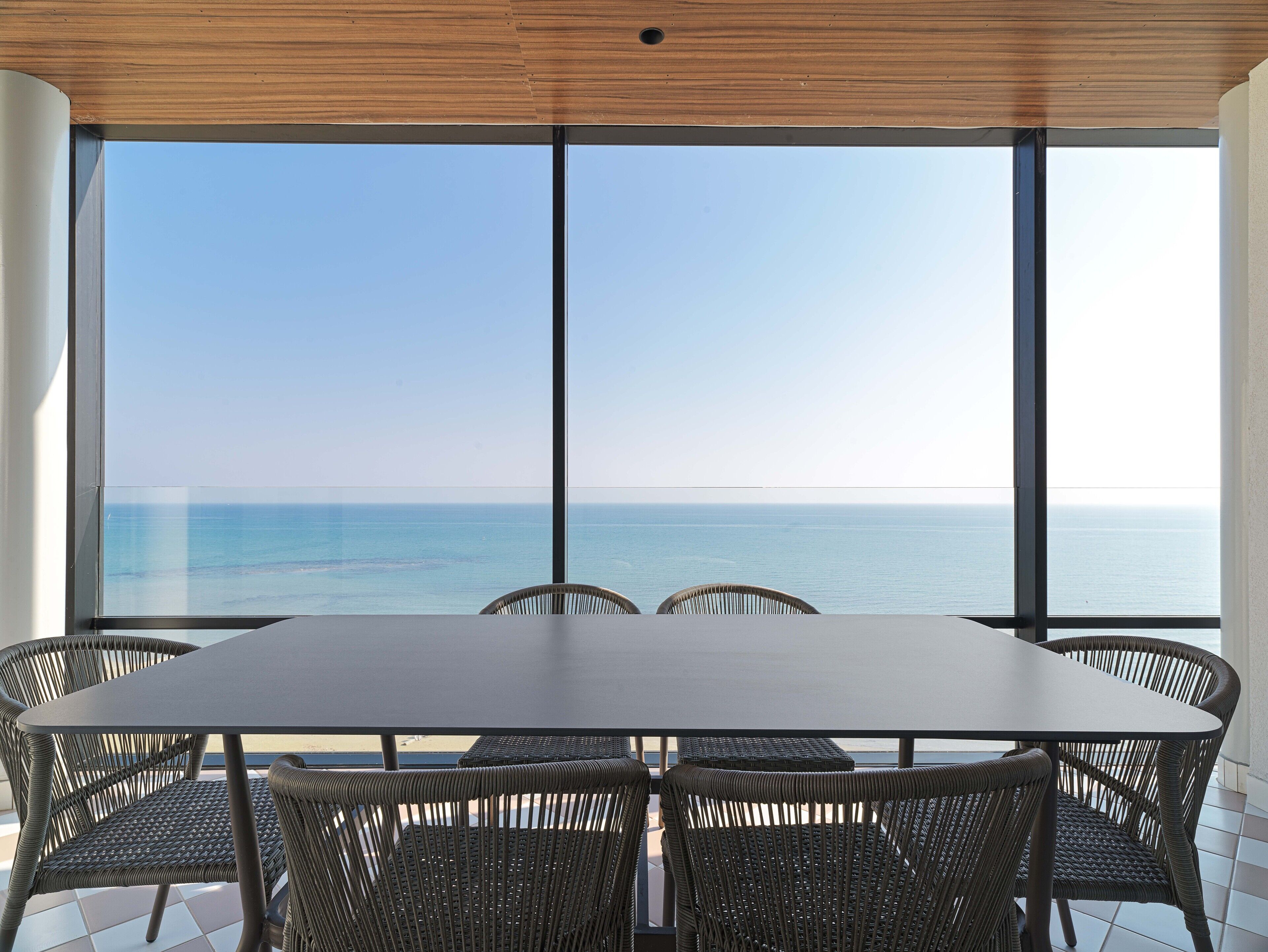 A dining area with a large table and wicker chairs, overlooking a calm sea.