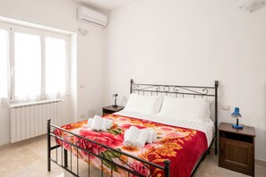 Cama bem arrumada com colcha florida e toalhas dobradas, mesa de cabeceira com abajur e janela com persianas.