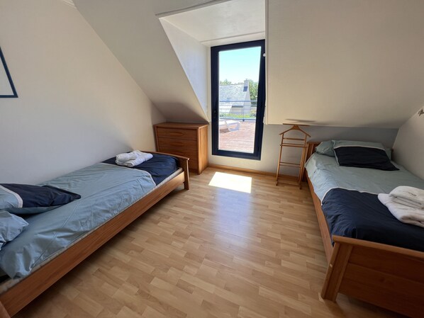 A small, well-lit room with two wooden beds, a wooden dresser, and a large window providing a view of the outside.
