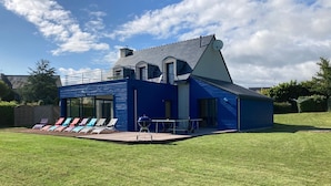 Una casa moderna de color blau amb un gran terrat, mobiliari exterior i zona de barbacoa.