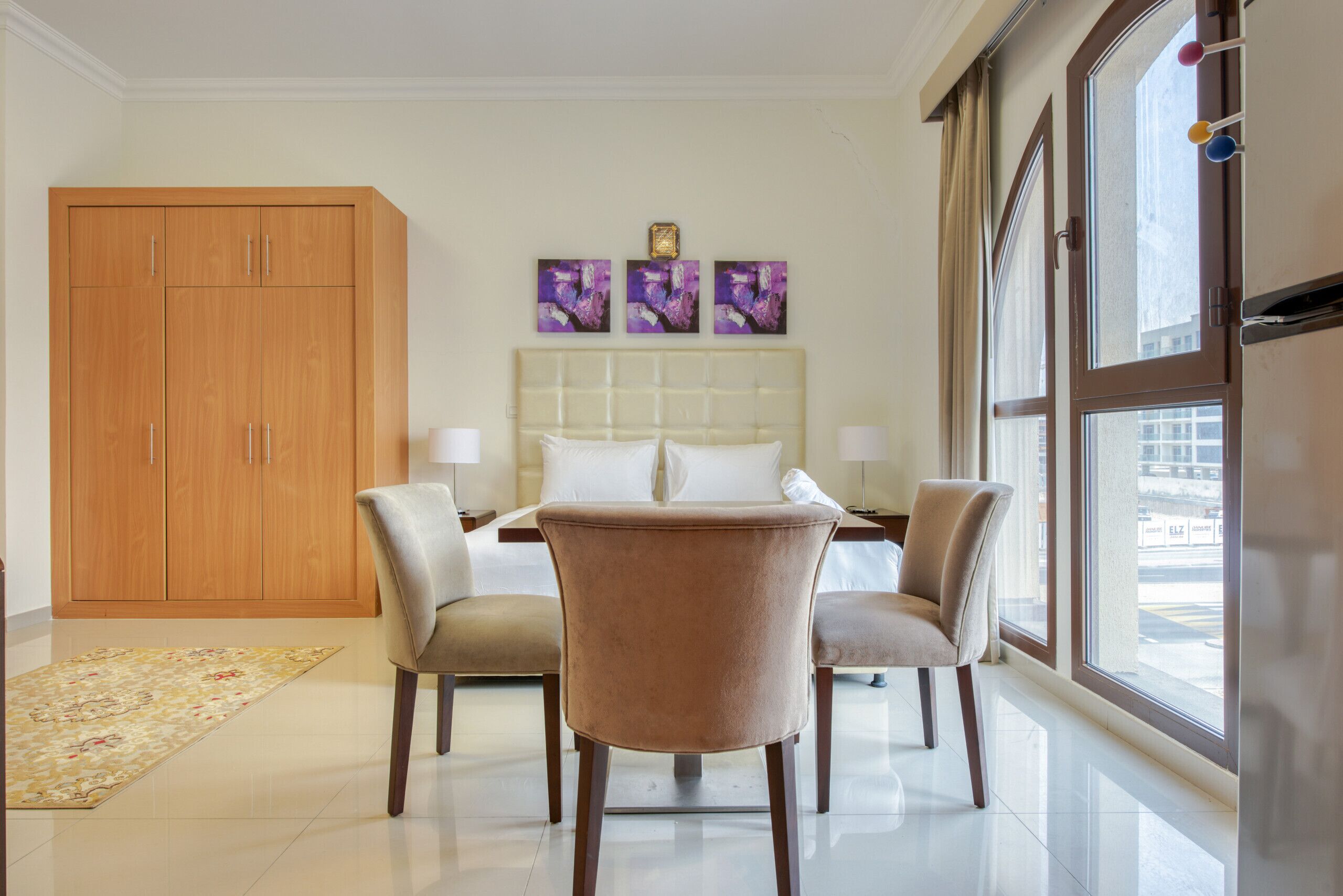 Habitación de hotel con una cama grande, dos mesitas de noche, un armario de madera y vistas a la ciudad a través de amplios ventanales.