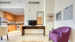 Een moderne woonkamer met een paarse fauteuil, een houten bureau, een flatscreen televisie en een keuken met houten kasten en een wasmachine.