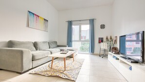 Un moderno salón con un sofá gris, una mesa de centro blanca, una alfombra estampada, un televisor sobre un mueble blanco y una ventana con cortinas azules.