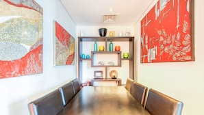 Un comedor con una mesa larga y sillas acolchadas, una estantería con jarrones coloridos y arte abstracto en las paredes.
