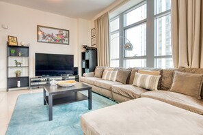 A modern living room with a large flat-screen TV, a beige sofa, a coffee table, and a large window with curtains.
