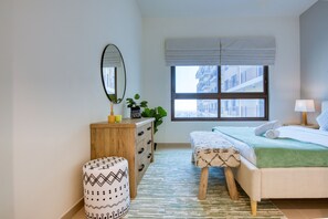 Une chambre à coucher avec un lit, une commode en bois, un miroir, une fenêtre donnant sur des immeubles et un tapis vert.