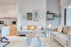 Un salon moderne avec un canapé gris, une table basse ronde et un tapis à motifs. On peut voir des reproductions botaniques encadrées au mur, ainsi qu’une petite table d’appoint avec une lampe.