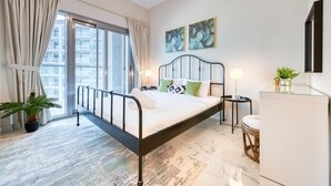 A modern bedroom with a bed, bedside tables, a desk, a potted plant, and a view of the city through large windows. - Exceptional 1BR in MAG 5, Emaar South by Deluxe Holiday Homes (Dubai)