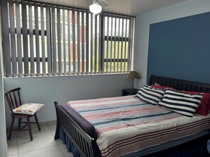 A bedroom with a bed, a chair, a nightstand, a lamp, and a window with blinds. - Family-friendly, sea and breaker view, 2 min from beach (Amanzimtoti)
