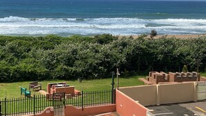 Une vue sur l’océan, avec les vagues qui déferlent sur la plage, une zone herbeuse bordée d’une clôture et un parking.