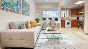 Ein Wohnzimmer mit einer Couch, Sesseln und einem Couchtisch. Es gibt eine Küchenzeile mit Schränken und Geräten.