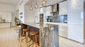 Una cocina moderna con una isla de mármol, taburetes de bar, luces colgantes y electrodomésticos de acero inoxidable.