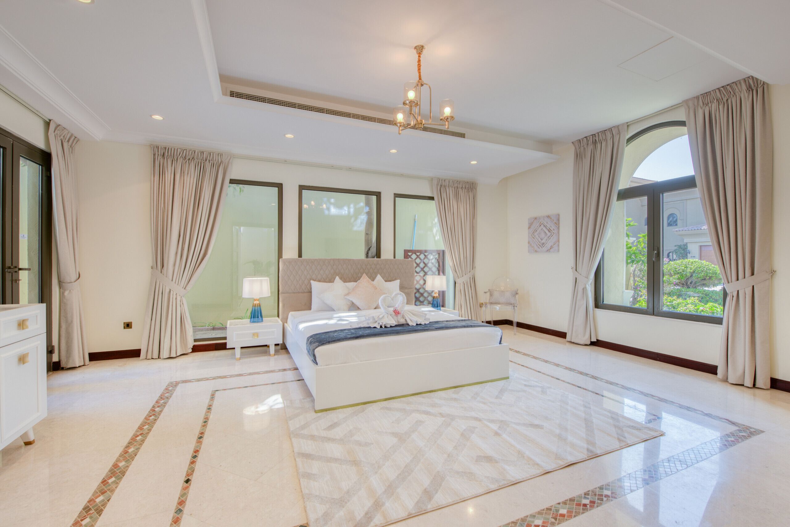 A spacious bedroom with a large bed, a nightstand, a lamp, and a view of the outdoors through large windows.
