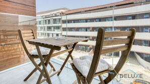 Varanda com mesa e cadeiras de madeira, vista para um edifício moderno com vários andares e grandes janelas.