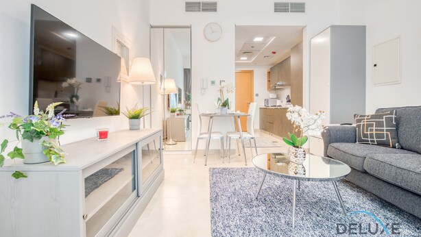 Uma sala de estar moderna com um sofá cinzento, uma mesa de centro e uma televisão. Há uma área de refeições com uma mesa e cadeiras.