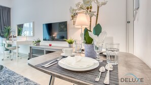 Uma área de jantar moderna com uma mesa posta para dois, incluindo pratos, talheres e copos. Há uma planta em vaso e um abajur na mesa.