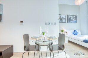 Une pièce moderne et bien éclairée, avec une table à manger en verre, deux chaises, un lit avec un oreiller bleu et un canapé. Des tableaux encadrés ornent le mur et une petite plante est posée sur la table.