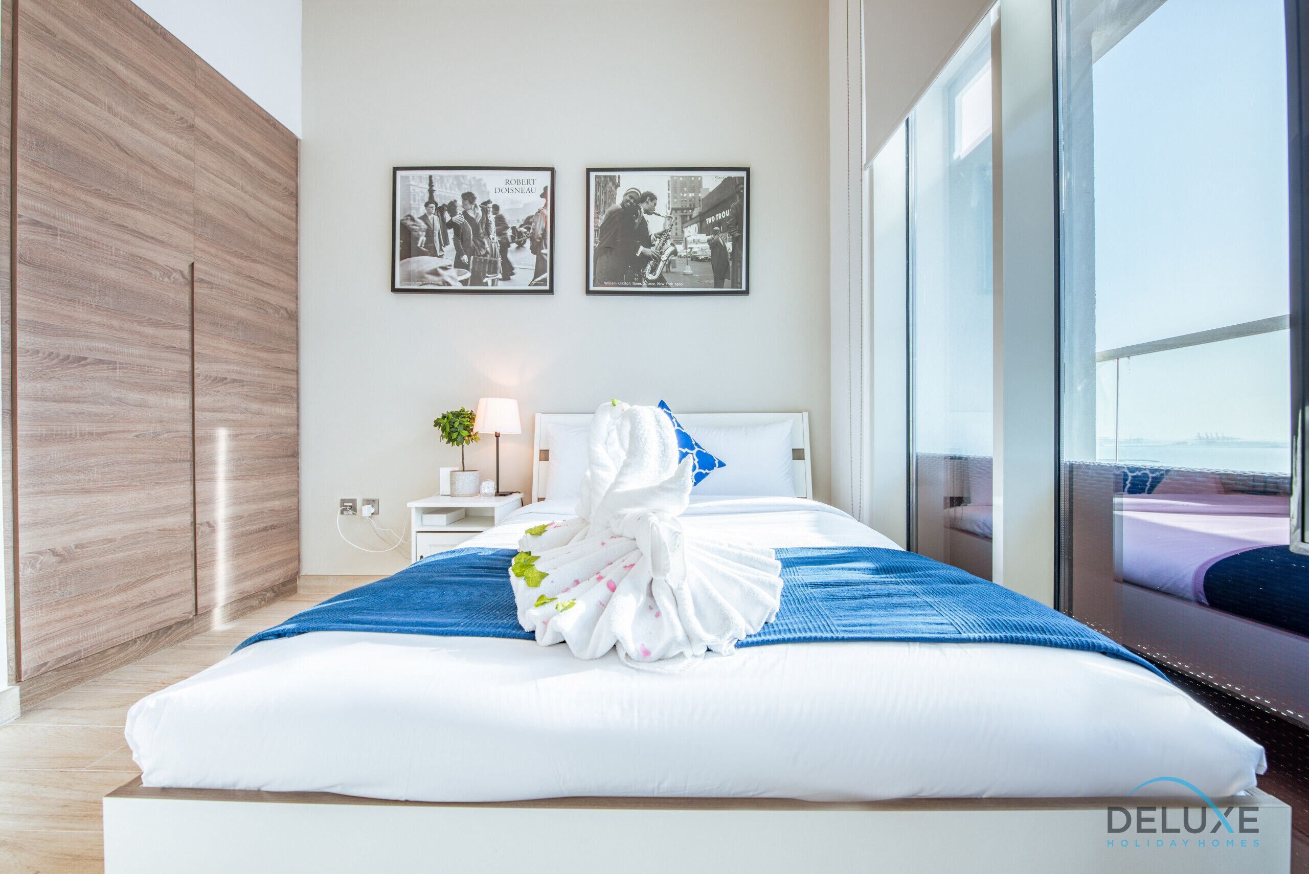A modern hotel room with a large bed, a wooden wardrobe, a nightstand with a lamp, and two framed pictures on the wall.