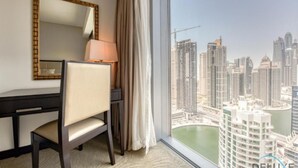 A hotel room with a desk, chair, lamp, and a large window offering a view of a cityscape with high-rise buildings and a body of water.