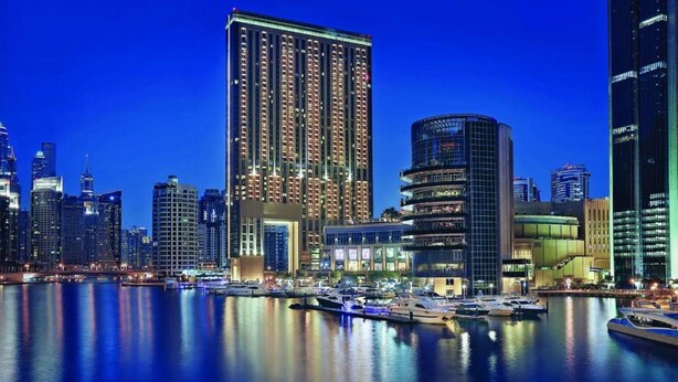 Een stadsbeeld bij nacht met moderne wolkenkrabbers, een jachthaven met boten en een opvallend hotel met een bijzondere, gebogen gevel.