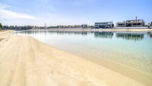 A beach with clear water, a sandy shore, and modern buildings in the background. - Signature 6BR Villa with Assistant Room and Private Pool in Frond F Palm Jumeirah by Deluxe Holiday Homes (Dubai)