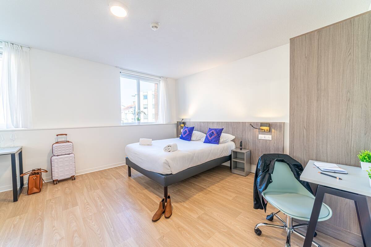 Ein modernes Hotelzimmer mit einem großen Bett, einem Schreibtisch und einem Stuhl. Auf dem Boden liegen ein Koffer und ein Paar Schuhe.