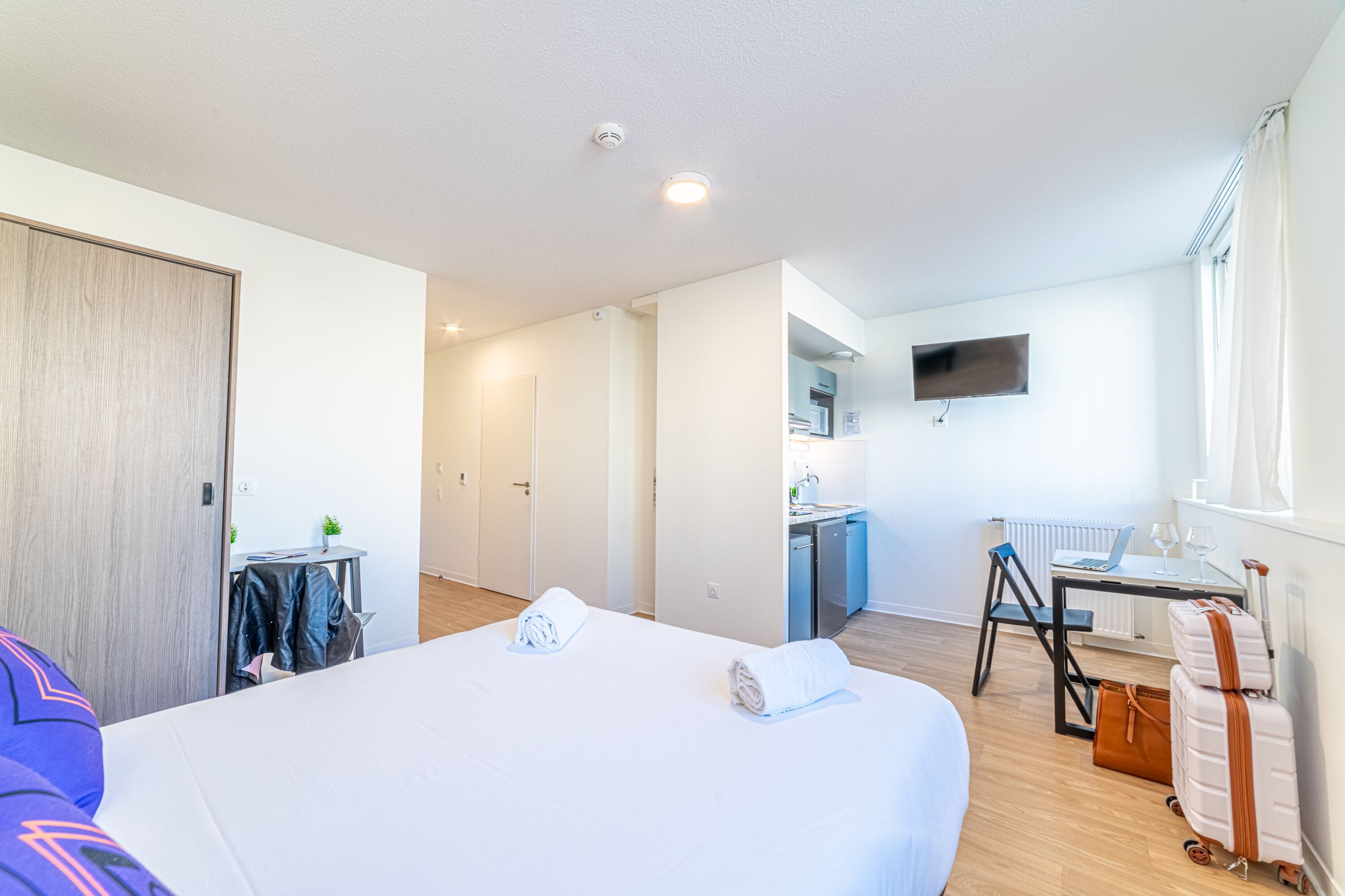 Habitación de hotel con cama, escritorio con silla, una pequeña kitchenette y televisor montado en la pared.