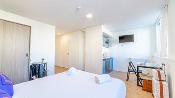 Habitación de hotel con cama, escritorio con silla, una pequeña kitchenette y televisor montado en la pared.