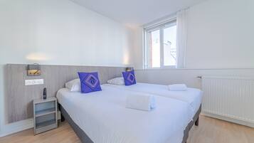 Habitación de hotel con dos camas, una mesita de noche, una lámpara y una ventana con cortinas.