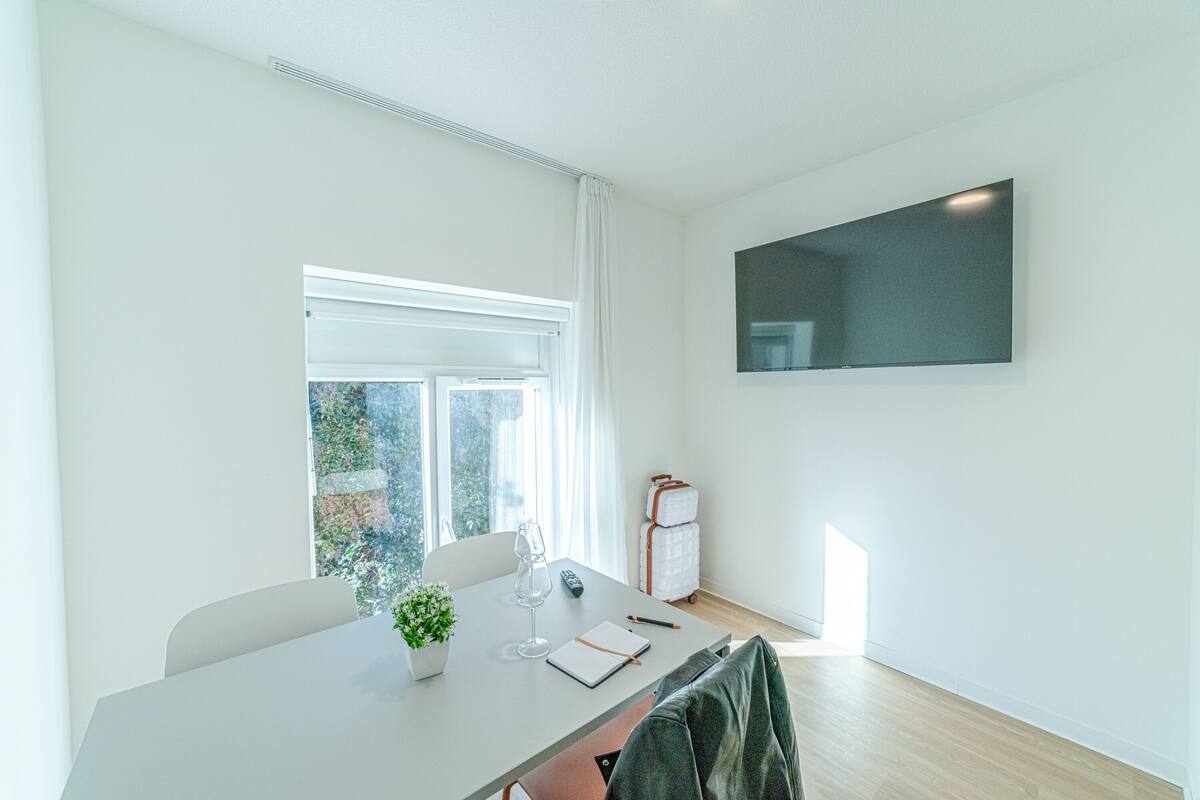 Ein modernes, gut beleuchtetes Zimmer mit einem großen Fenster, einem an der Wand befestigten Flachbildfernseher, einem Tisch mit Notizbuch, Stift und einem Glas Wasser sowie einem Koffer in der Ecke.