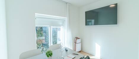 A modern, well-lit room with a large window, a flat-screen TV mounted on the wall, a table with a notebook, pen, and a glass of water, and a suitcase in the corner.
