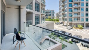 Um prédio de apartamentos moderno com varanda, que possui guarda-corpo de vidro, área de estar com duas cadeiras e vista para um estacionamento com vários carros.