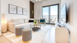 A modern living room with a sofa, ottomans, a coffee table, and a TV. There are framed artworks on the wall and a balcony with a view. - Spotless 1BR at UNA Town Square Dubailand by Deluxe Holiday Homes (Dubai)