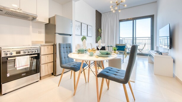 A modern kitchen with a built-in oven, a dining area with a round table and blue chairs, and a large window with a view of the city. - Spotless 1BR at UNA Town Square Dubailand by Deluxe Holiday Homes (Dubai)