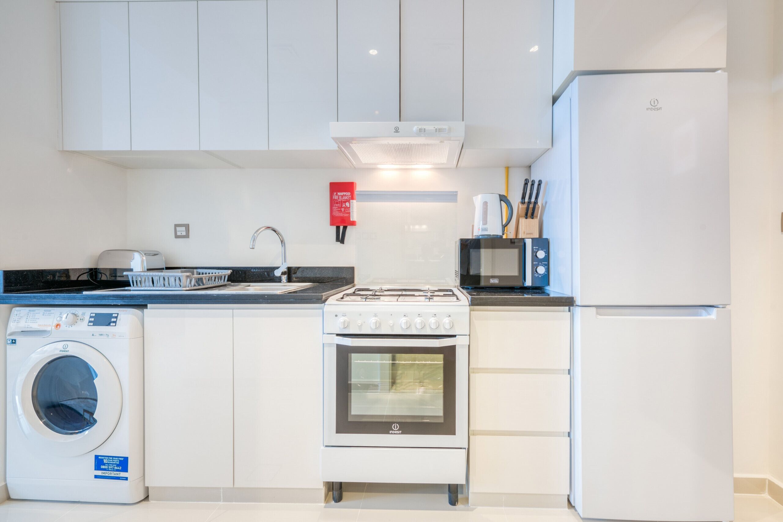 A modern kitchen with white cabinetry, a built-in oven, a microwave, a washing machine, and a coffee maker.