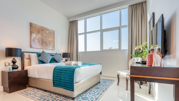 A modern hotel room with a large bed, a desk, a chair, a TV, and a window with curtains. - Intimate Studio in Damac Ghalia Jumeirah Village Circle by Deluxe Holiday Homes (Dubai)