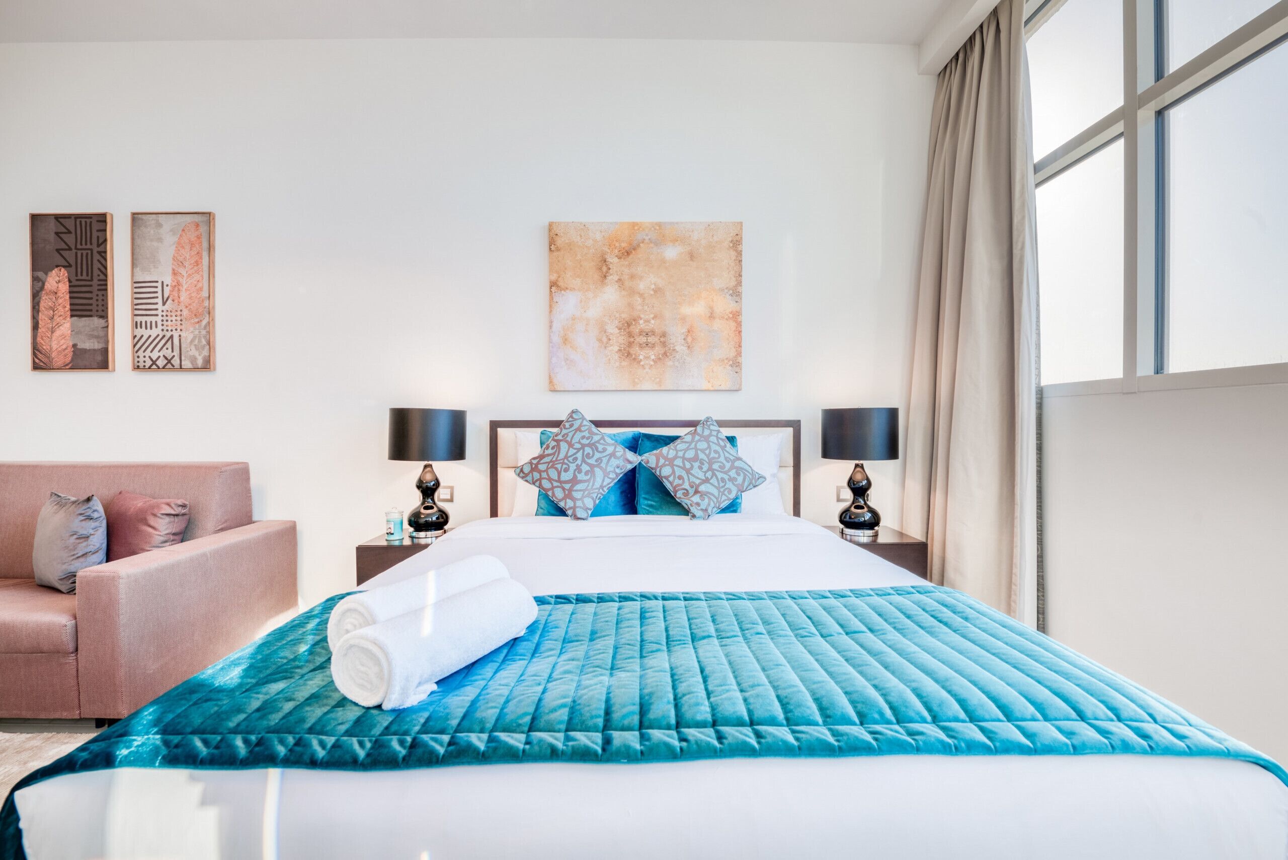 A hotel room with a bed, a sofa, two wall art pieces, and a window with curtains.